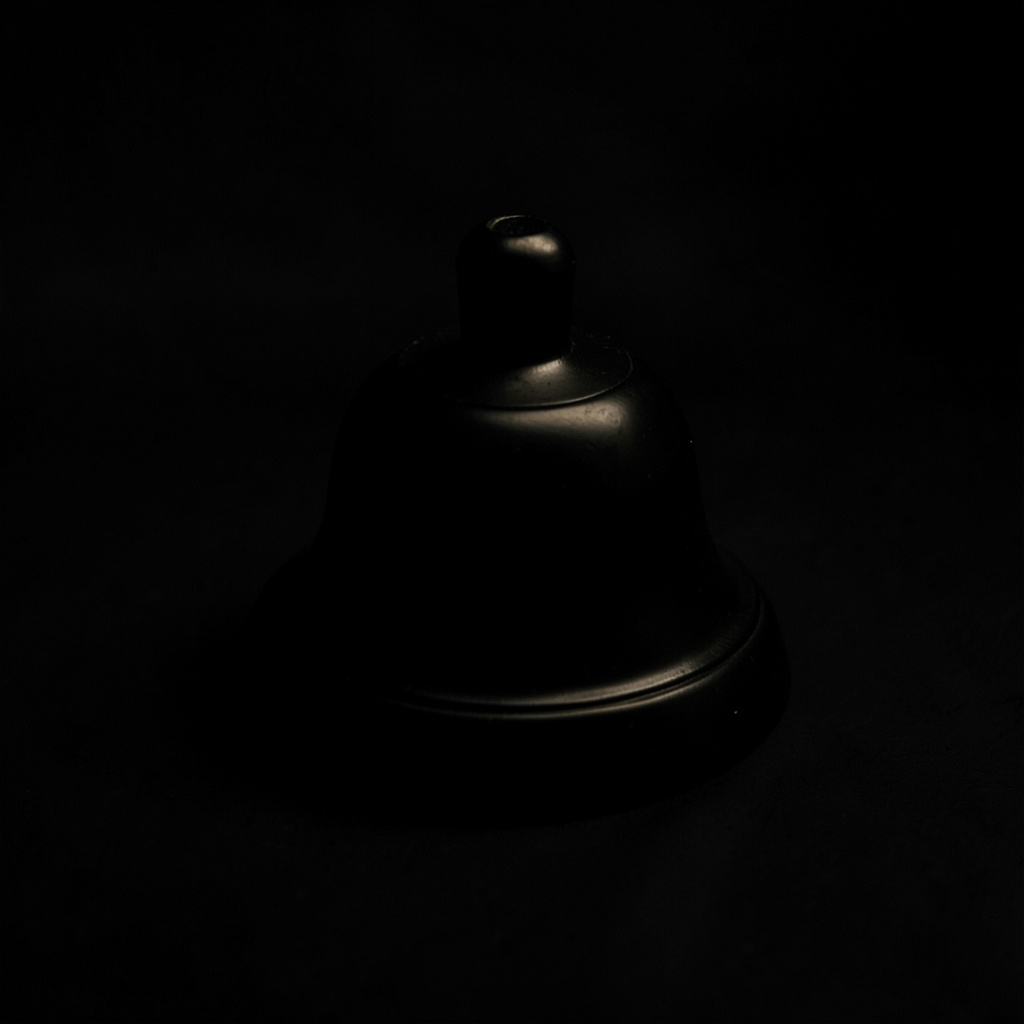 Dark low-key photograph of a softly lit help-desk bell on a matte black surface, deep shadows pooling around the base.